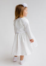 Lade das Bild in den Galerie-Viewer, Das gleiche Mädchen mit dem weißen Babydirndl von Anna Kaval, mit Bolero und einem süßen Blumenhaarband, dreht sich fröhlich um und zeigt das elegante Taufkleid von hinten.