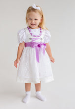 Lade das Bild in den Galerie-Viewer, Fröhliches Kind in einem Babydirndl von Anna Kaval. Das Flieder Taufdirndl kombiniert zarte Spitze und eine schöne Schürze, ideal für festliche Anlässe.