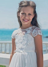 Lade das Bild in den Galerie-Viewer, Vergnügliches Blümenmädchen im Kommunionskleid von Anna Kaval, mit zarter Spitze und einem strahlenden Lächeln, vor einer malerischen Meereskulisse.