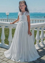 Lade das Bild in den Galerie-Viewer, Ein Mädchen in einem langen, eleganten Blümenmädchen Kommunionskleid aus Spitze von Anna Kaval, lächelnd am Ufer mit Meer im Hintergrund.
